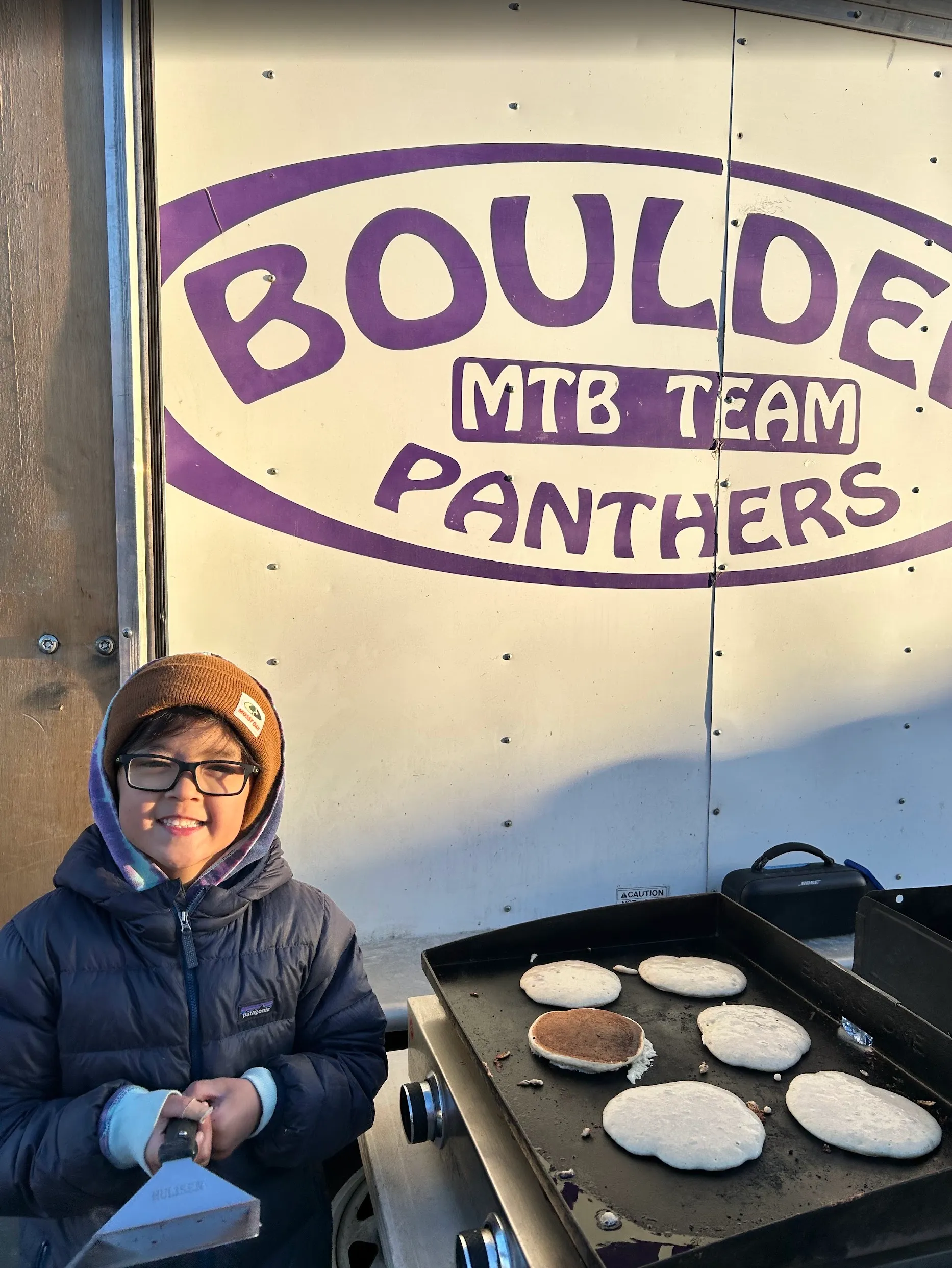 A young rider smiling in front of the Boulder MTB Team Panthers trailer, with pancakes cooking on a griddle nearby